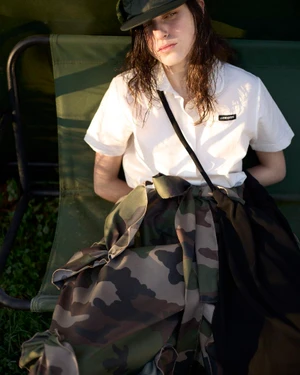 Model Margaux Martin seated outdoors in a white polo shirt, dark cap, and camouflage print fabric draped across her lap.