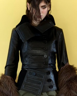 Model Margaux Martin in a structured black leather buttoned jacket with fur trim against a yellow studio backdrop.