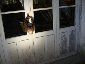 Brown fringe hairpiece accessory hanging from the latch of a weathered white glass-paneled door in direct sunlight.