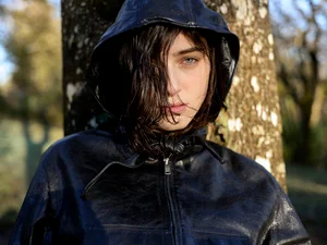 Close-up of model Margaux Martin wearing a black hooded leather jacket leaning against a tree trunk in dappled sunlight.