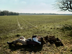 Model Margaux Martin lying on grass beside chopped firewood in an expansive open rural field under bright natural daylight.