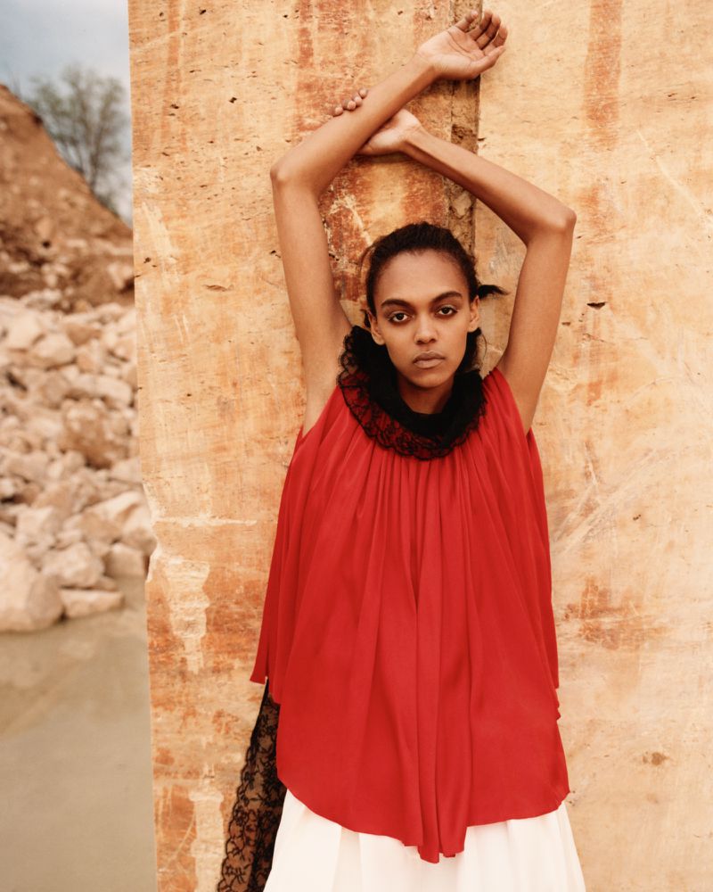 Model with arms raised posing against a textured stone wall in an outdoor desert or quarry setting