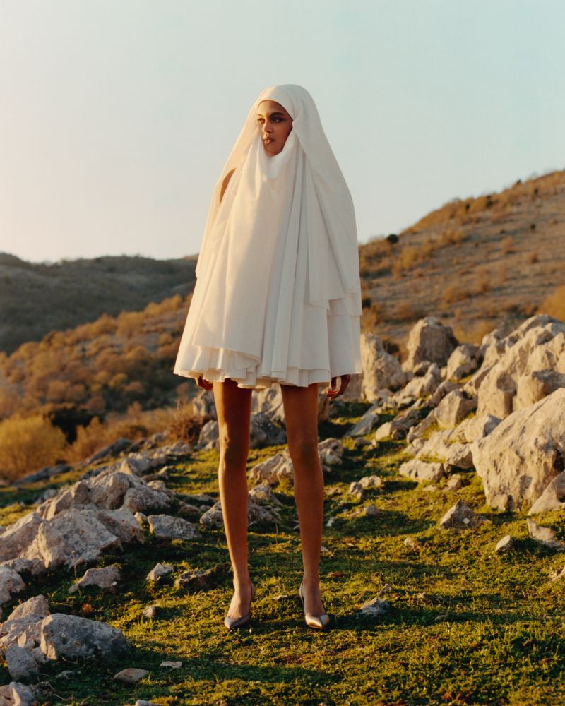 Model in a white draped hooded dress standing outdoors on a rocky hillside at sunset