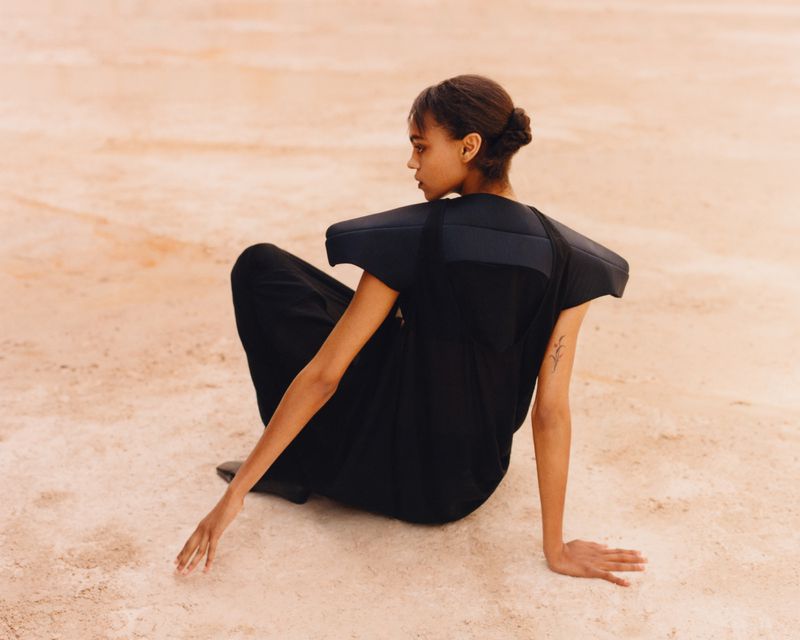 Model seated on pale sand wearing a black structured garment with broad padded shoulders