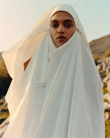 Model draped in white fabric standing outdoors against a rocky landscape under a soft sky for Present Space.