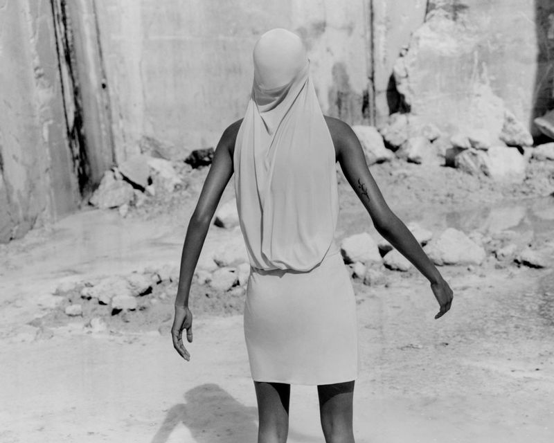 Model wearing a light-coloured cowl neck dress and head covering standing with arms out against a rocky backdrop.