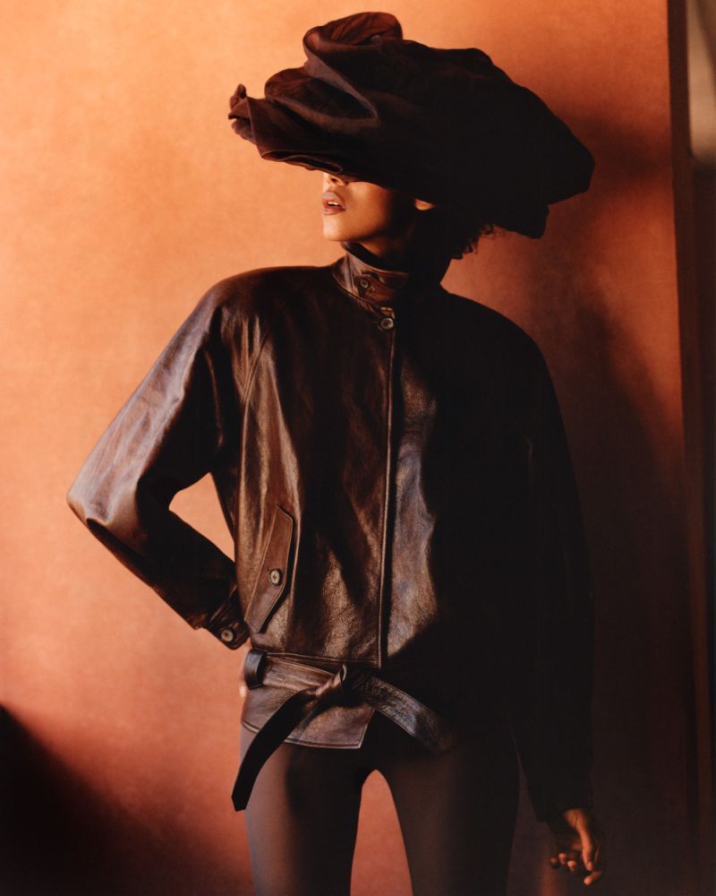 Model wearing dark leather jacket and sculptural dark headpiece against a warm terracotta background.