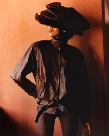 Model wearing dark leather jacket and sculptural dark headpiece against a warm terracotta background.