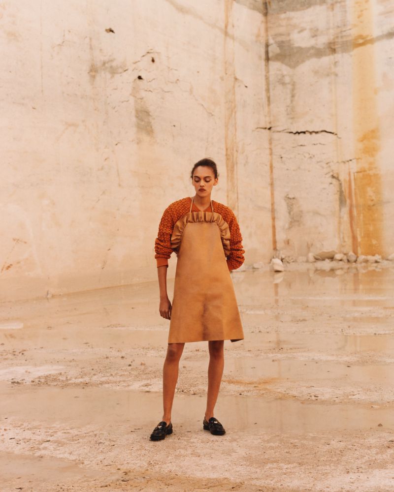 Model standing in textured apron dress against a weathered stone quarry wall with shallow water on the ground.