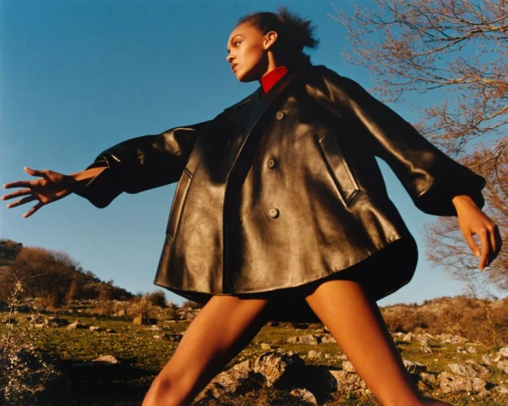Model wearing a black leather jacket over a red top outdoors against a blue sky and grassy hillside.