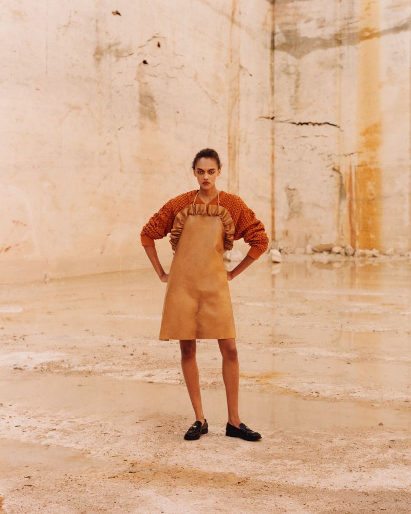 Model standing in a textured quarry wearing an orange sweater and tan apron dress with loafers