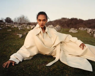 Model lying on grass wearing a long cream coat in a rocky landscape