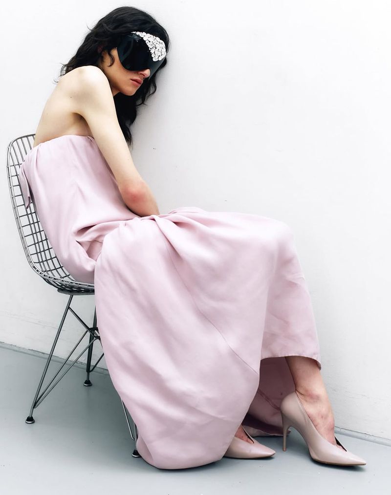 Elegant woman in pink gown sits on wire chair against white wall, wearing a black bejeweled eye mask and matching high heels.