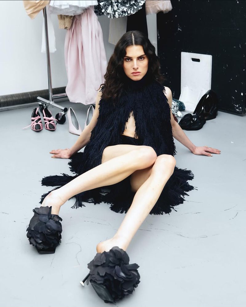 Model Lou Seriot sitting on a grey floor wearing a black fringed top for Exhibition Magazine