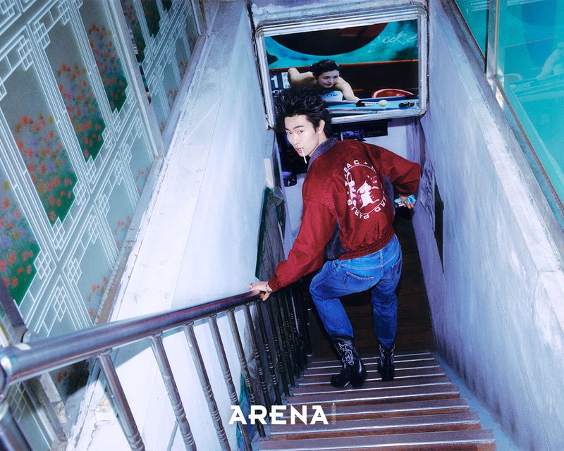 Man in a red bomber jacket and blue jeans walking down a narrow concrete indoor stairway holding a handrail