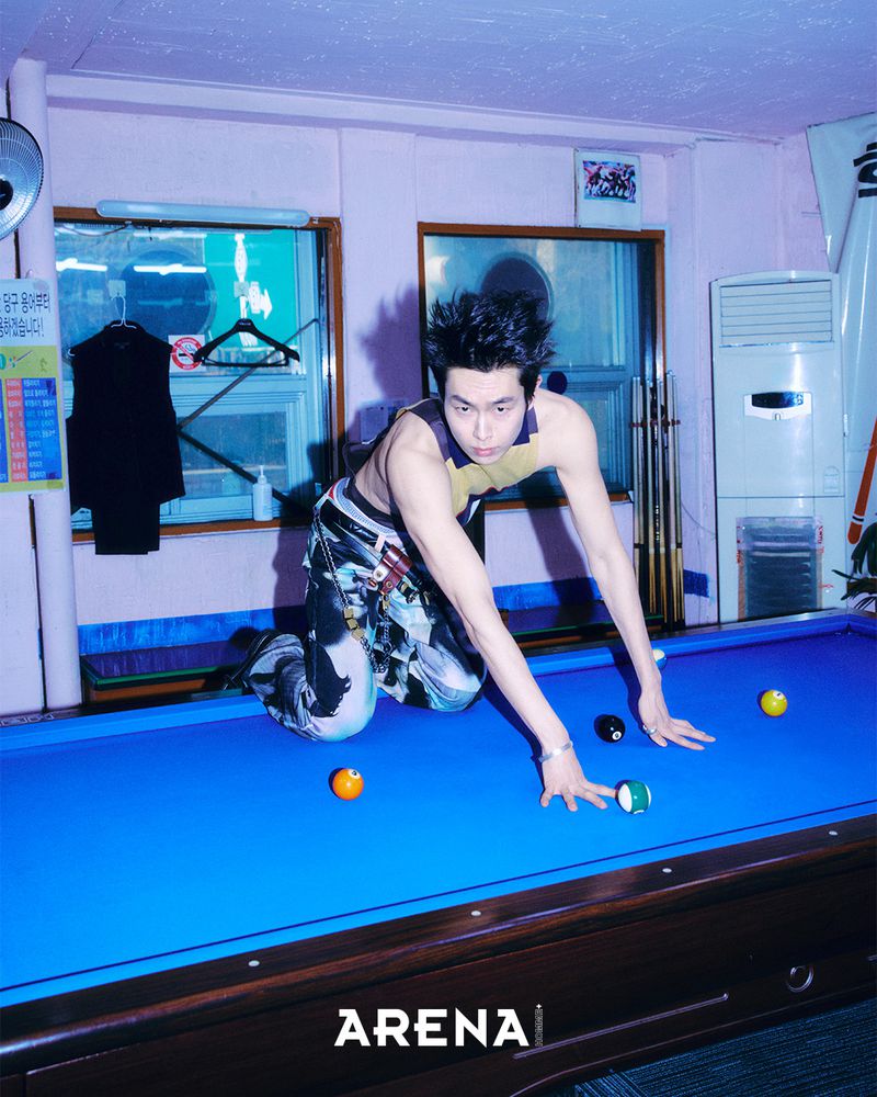 A man kneeling on a blue pool table indoors in a room with pink walls, wearing patterned trousers and a sleeveless top.