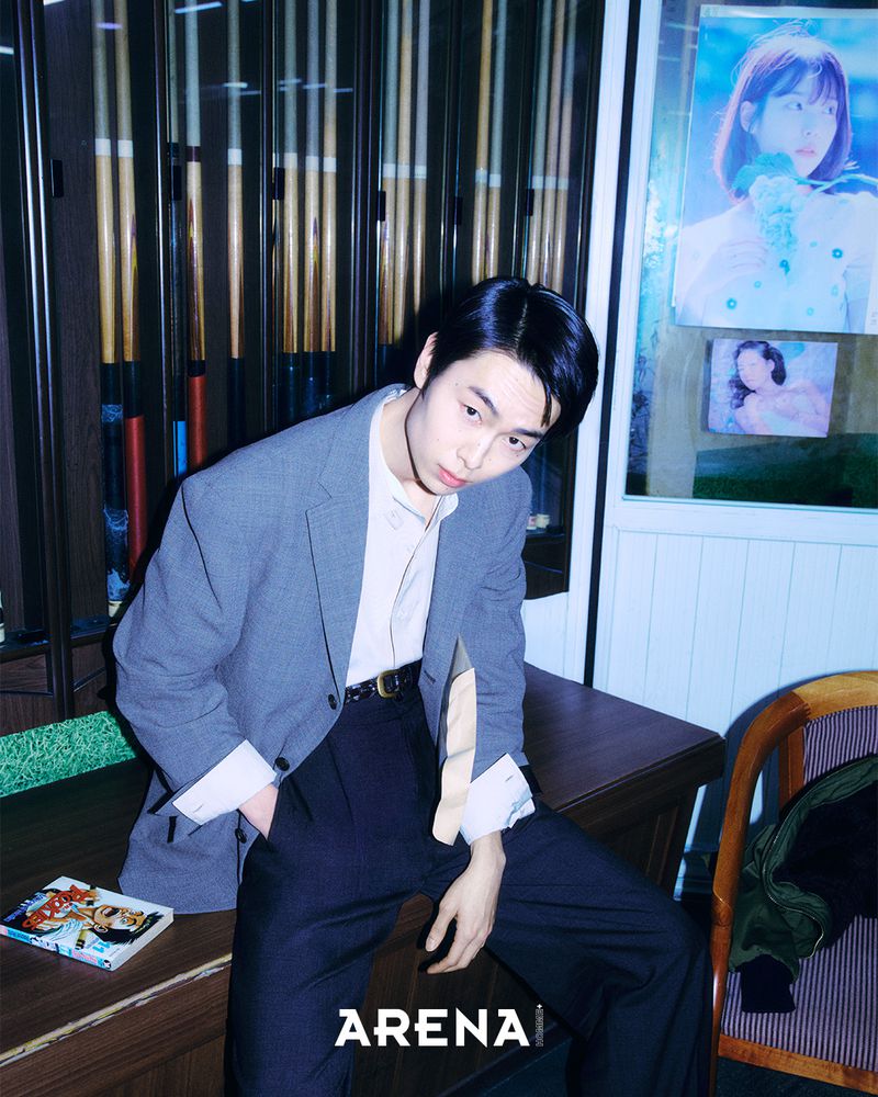 Man wearing a grey blazer and dark trousers seated on a desk in an interior room with a poster on the wall