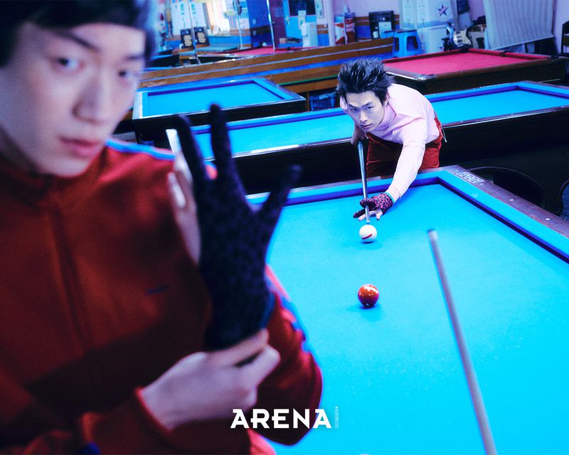Two men playing billiards in a pool hall, with one in the foreground wearing a red track jacket adjusting a black glove.