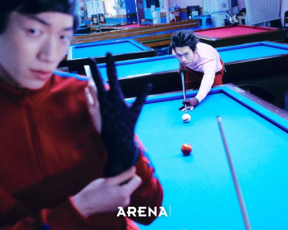 Two men playing billiards in a pool hall, with one in the foreground wearing a red track jacket adjusting a black glove.