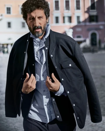 Man with curly dark hair and beard wearing a black jacket and striped shirt outdoors in a public square