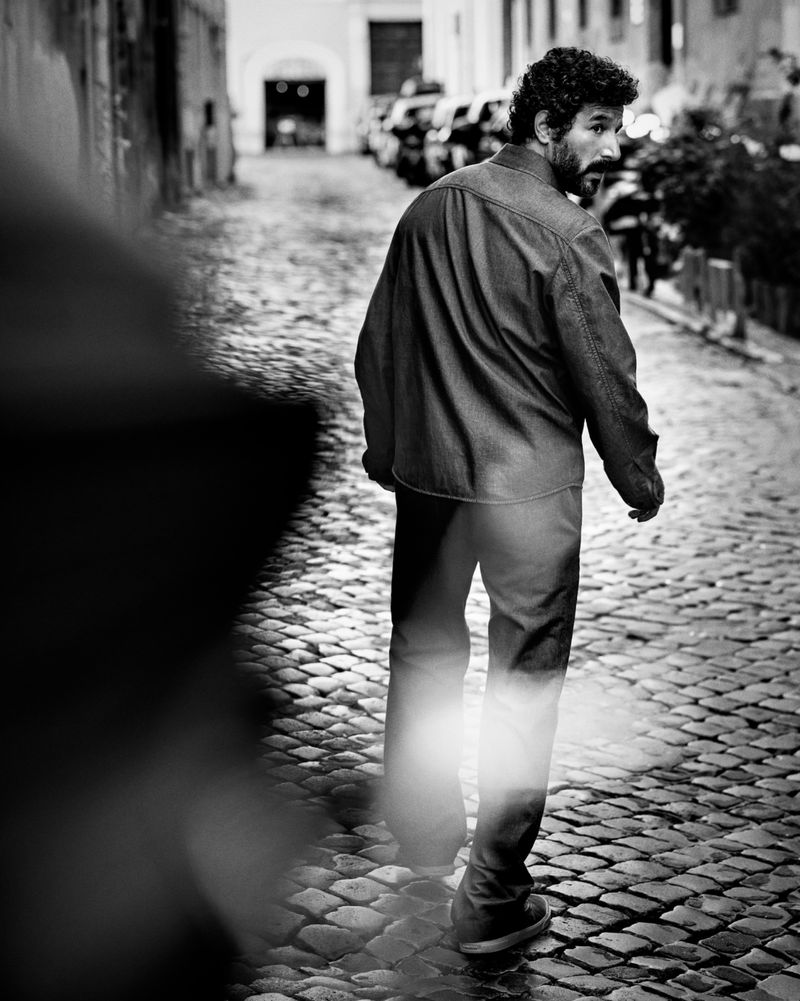 Man with curly hair and beard walking down a cobblestone street looking back over his shoulder for Moncler collection campaign.