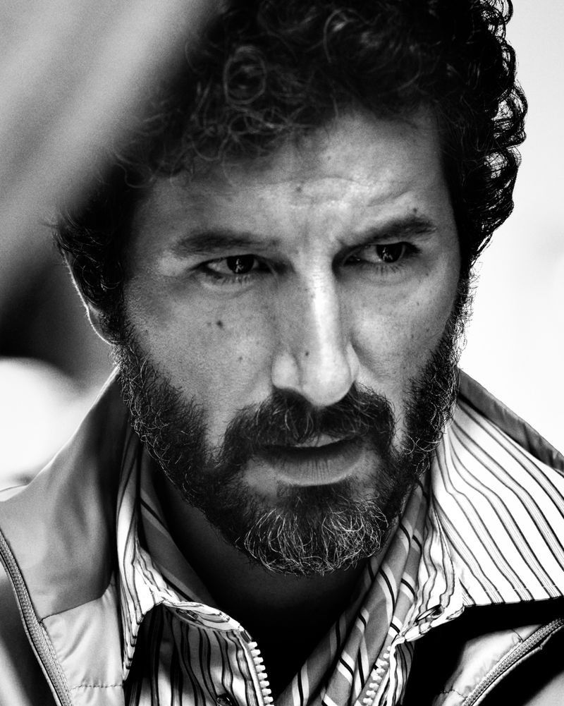 Close-up monochrome portrait of a man with curly hair and a beard wearing a striped shirt and jacket for Moncler.