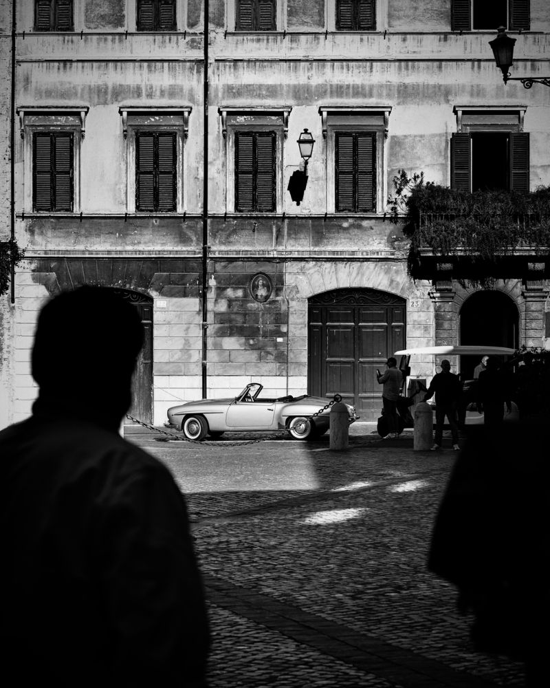 Monochrome street scene featuring a vintage convertible parked in front of an old European building with shuttered windows.