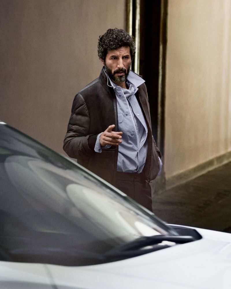 Man wearing a brown jacket standing outdoors near a car with an urban wall background