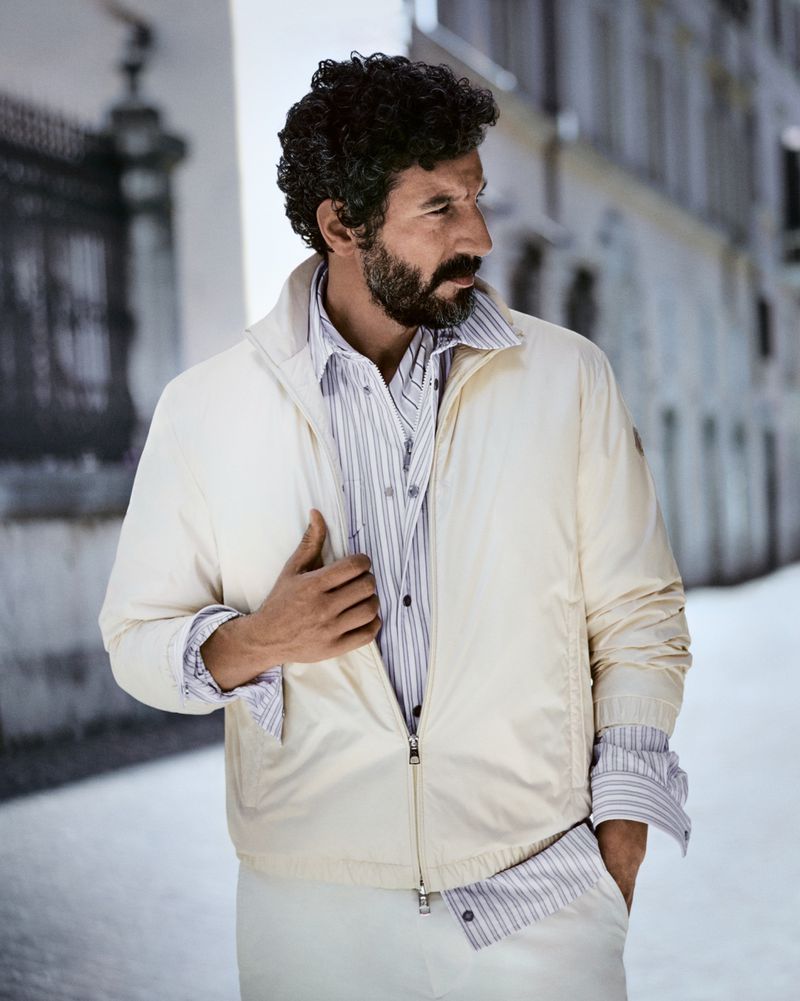 Man with dark curly hair and beard wearing an off-white jacket and striped shirt standing outdoors on a street