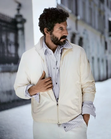 Man with dark curly hair and beard wearing an off-white jacket and striped shirt standing outdoors on a street