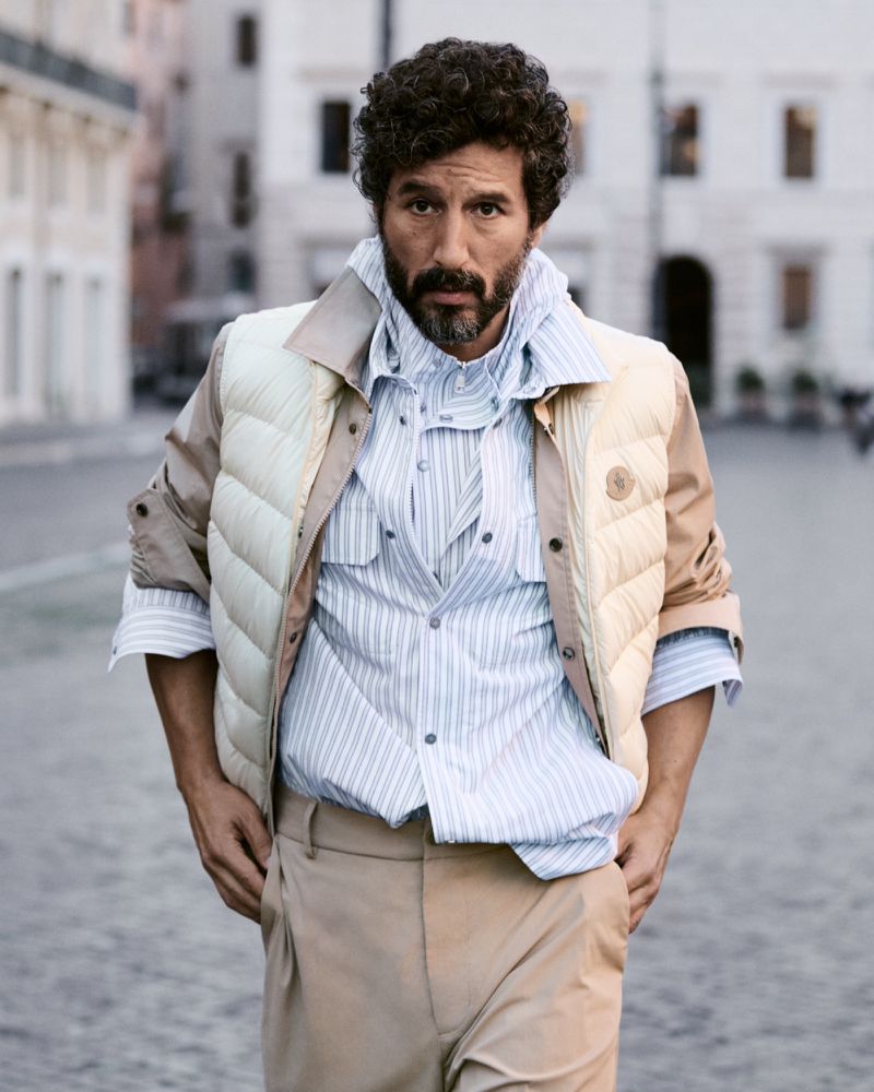 Man with curly dark hair and beard wearing a striped shirt and puffer vest outdoors on a city street.