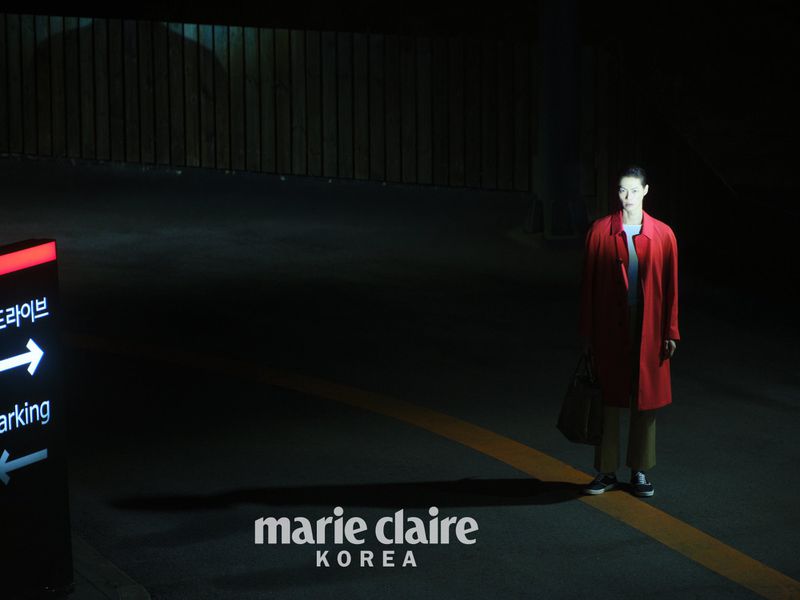 Model standing outdoors at night in a bright red coat and trousers under harsh spotlight, holding a handbag.