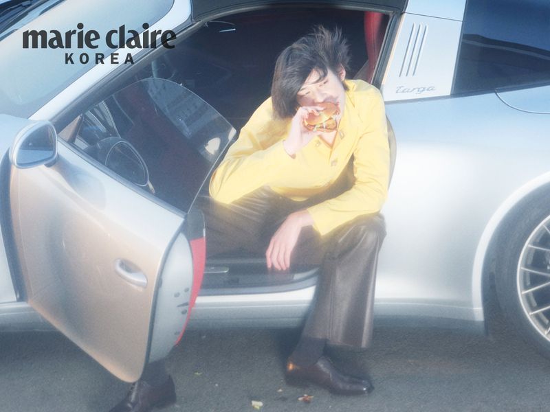 Model in a yellow top and dark trousers seated inside a silver sports car eating a burger