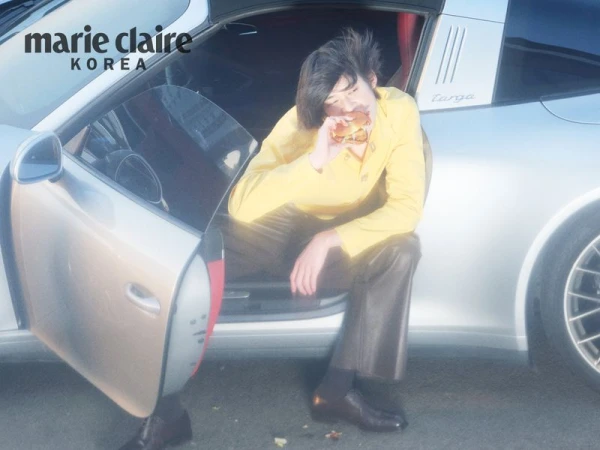 Model in a yellow top and dark trousers seated inside a silver sports car eating a burger