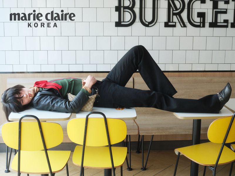Model lying across restaurant tables wearing a black jacket and pants with yellow chairs in the foreground.