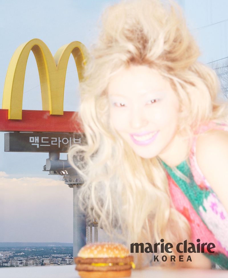 Blonde model smiling with a burger and a McDonald's sign in the background for Marie Claire Korea editorial.