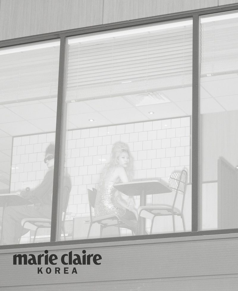 Models seated inside a restaurant viewed through large exterior windows in a black and white editorial photograph