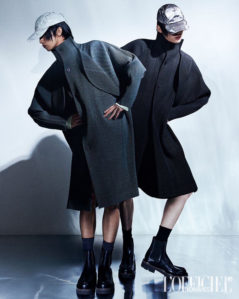 Two male models in pleated coats and metallic caps posing against a grey studio backdrop for L'Officiel Korea Duo project