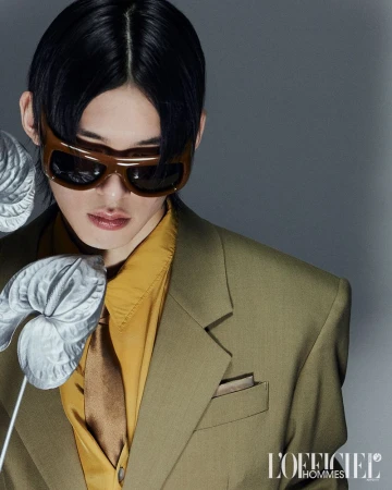 Model wearing a structured olive blazer and sunglasses posing beside metallic botanical stems against a grey studio backdrop.