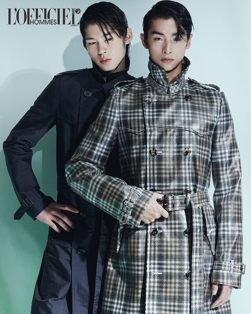 Two male models posing against a light background for L'Officiel Korea, wearing dark trench coats.