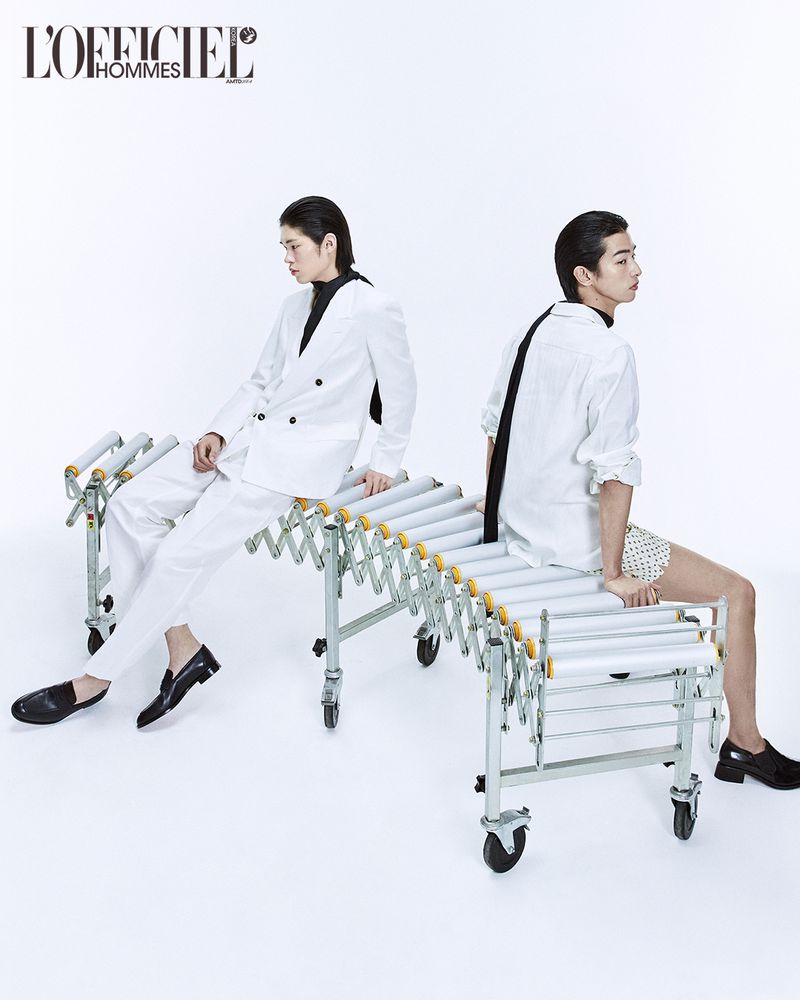 Two male models in white outfits seated back-to-back on an industrial conveyor roller bench against a plain white background.