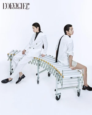 Two male models in white outfits seated back-to-back on an industrial conveyor roller bench against a plain white background.