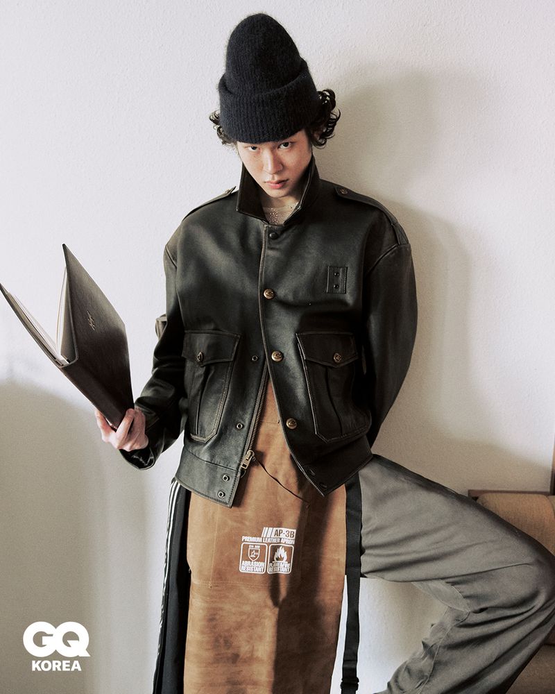Model in black beanie and dark leather jacket holding a dark book against a light wall for GQ Korea
