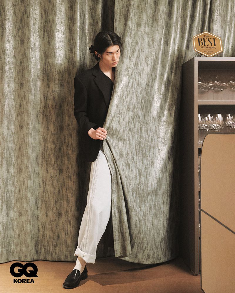 Model with long dark hair in a black blazer and white pants peeking through heavy textured curtains for GQ Korea editorial.
