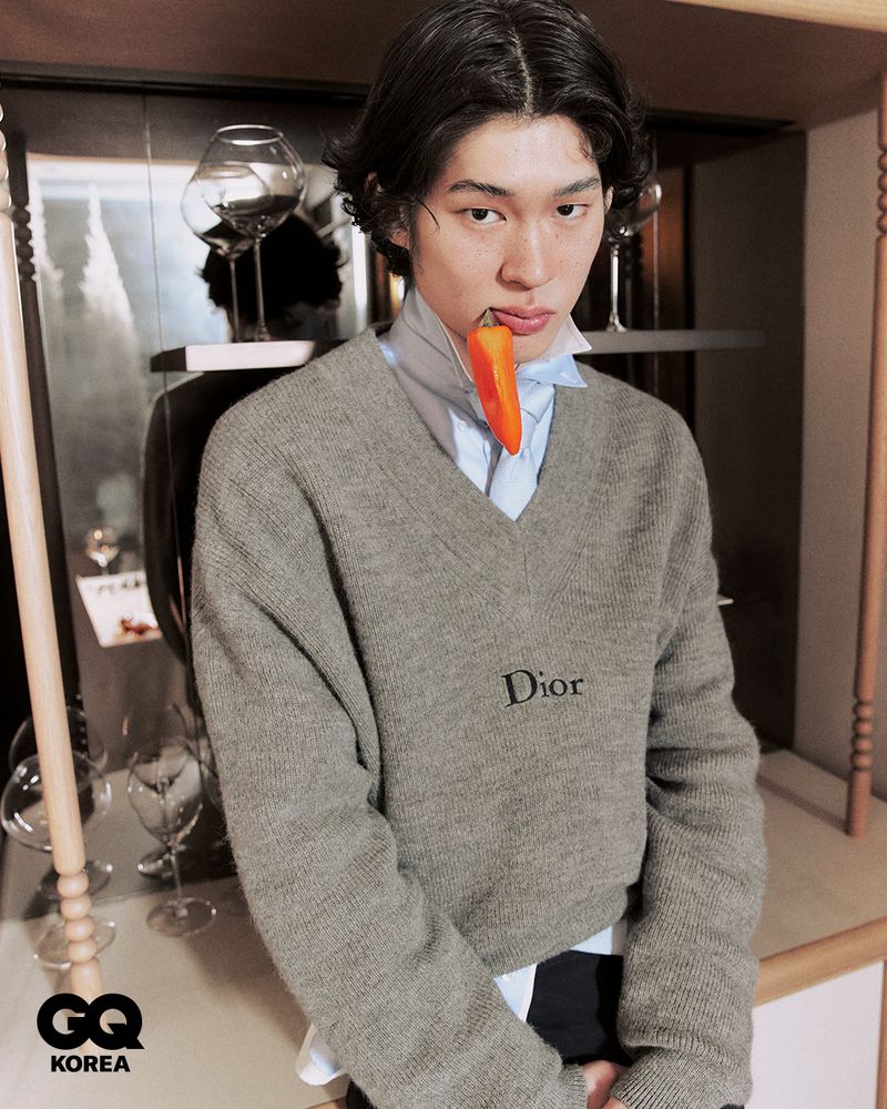 Model wearing a grey v-neck sweater biting an orange pepper for a GQ Korea fashion editorial.