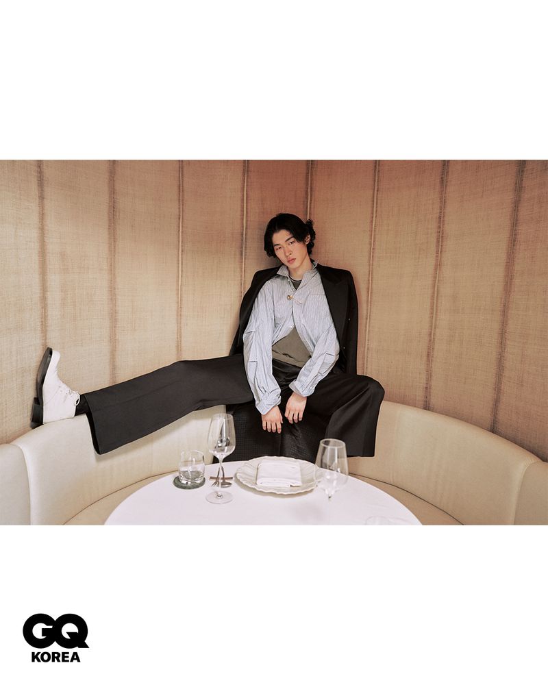 Model seated inside a restaurant booth with one foot propped up on the cushion, wearing a black blazer and wide trousers.
