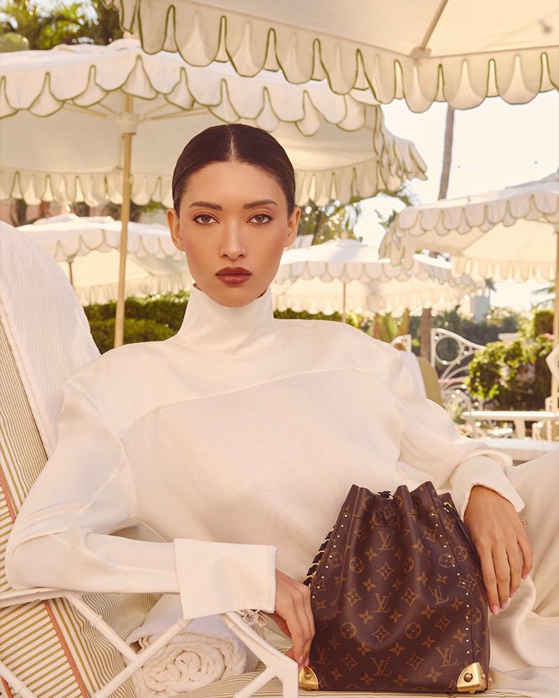 Model wearing a white high-neck top seated outdoors under white patio umbrellas with a monogram bucket bag.