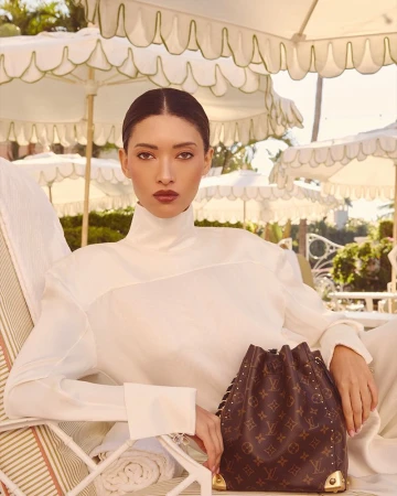 Model wearing a white high-neck top seated outdoors under white patio umbrellas with a monogram bucket bag.