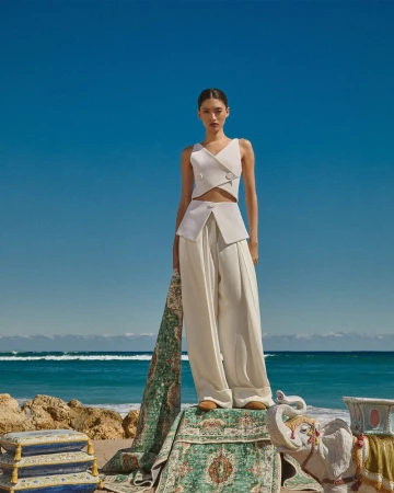 Model wearing white sleeveless top and wide-leg trousers standing on patterned green rug by ocean under clear blue sky.