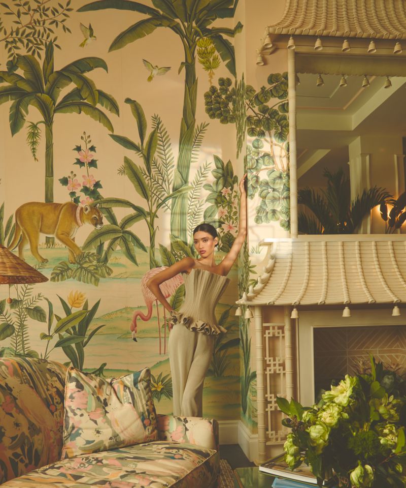 Model wearing a structured cream outfit poses against an ornate botanical wallpaper mural in an elegant interior setting.
